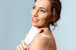 A beautiful smiling woman is holding a tube of moisturizer and sunscreen near her shoulders..