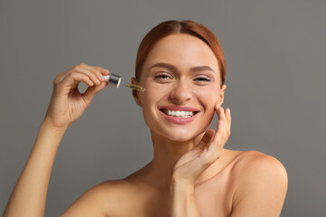 Beautiful young woman applying cosmetic serum onto her face on grey background