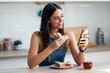 © nenetus - Beautiful sporty woman eating healthy sanwich while using smartphone in the kitchen at home