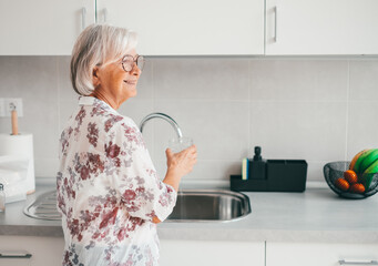 Smiling gray haired senior woman in home kitchen drinking tap water from glass