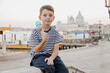 Сute seven-year-old boy in a striped t-shirt sits on the waterfront in Venice in the evening and eats gelato (ice cream)