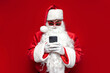 © Богдан Маліцький - santa claus in hat and festive glasses uses smartphone on red background, man in santa costume types online