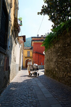 Beautiful Mother Pushing A Stroller With Sleeping Baby, Walking Along A Cobblestone Alley In Medieval Italian Canzo Town