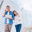 © Mallika - Portrait of cheerful couple with laptop and pointing finger in the copy space, Smiling couple Asian people standing over modern building background with copy space.