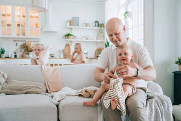 Careful father dressing little boy sitting on couch at home. Cute family moments. Child boy sitting on fathers knees while sisters at kitchen. Family care.