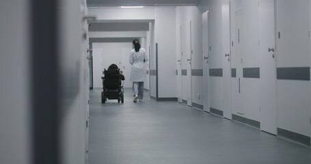 Wall Mural - Female doctor walks down the clinic hallway, consults woman with physical disability. Physician talks about medical procedures with patient in electric wheelchair. Corridor of modern medical facility.