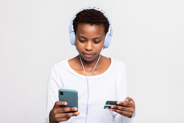 Serious concentrated dark skinned black woman holding credit card and cell phone in hands doing purchase shopping online wearing headphones earphones listening to music