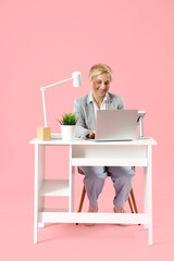 Mature female programmer working with laptop at table on pink background