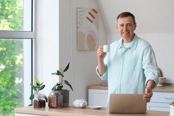 Wall Mural - Mature man with cup of coffee and laptop messaging in kitchen