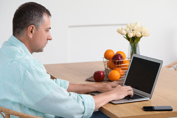 Wall Mural - Mature man with laptop messaging at table in kitchen