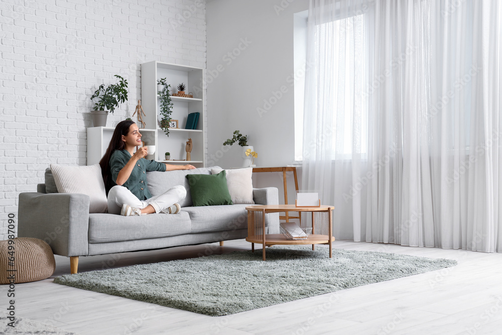 Beautiful young woman with cup of coffee sitting on sofa in living room ...