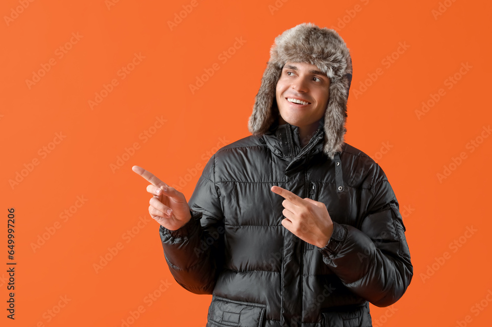 Young man in winter clothes pointing at something on orange background