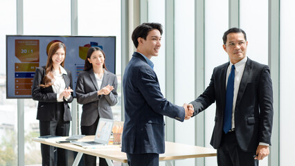 Asian professional successful male businessman in formal suit standing smiling shaking hands with client customer when female businesswoman colleague applauding clapping hands celebrate job deal done