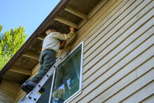 Senior Man On An Extension Ladder Replacing The Mesh Of An Attic Vent On The Exterior Of A Residential Building
