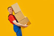 Laughing loader carries large boxes. Yellow background. Portrait of young man with pyramid of cardboard boxes in his hands. Copy space, mock up.