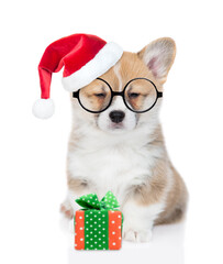 Sticker - Funny Pembroke welsh corgi puppy  wearing eyeglasses and red  christmas hat  sits with gift box and looks at camera. isolated on white background