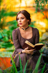 Wall Mural - Beautiful red haired girl in brown long medieval dress and black vintage choker sitting on stone wall. Art work of romantic lady with book.Pretty tenderness model posing on nature.