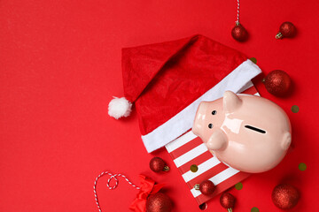 Wall Mural - Piggy bank on gift box, balls and Santa hat on red background, space for text