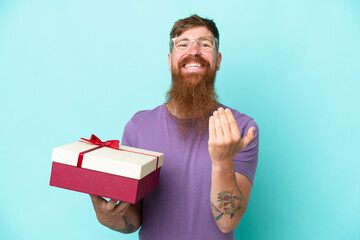 Wall Mural - Redhead man with long beard holding a gift isolated on blue background inviting to come with hand. Happy that you came