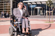 © Raul Mellado - smiling businesswoman using wheelchair talking by mobile phone in front of an office building in the financial district, concept of diversity and technology of communication
