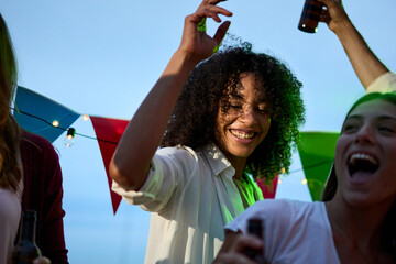 Happy young African adult afro woman dancing with friends at rooftop party. Excited people have fun drinking beer outdoor in dusk. Lively Millennial female celebrating summer vacation.