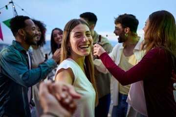 POV young smiling Caucasian woman holding hands boyfriend at party. Attractive girl looking cheerful on camera friends in background outdoors at dusk. People gathered on rooftop celebrating birthday.