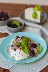 Wall Mural - Feta cheese sliced on blue plate with sesame, flax seeds, served with black olives and basil leaves. Delicious and healthy breakfast on terrace