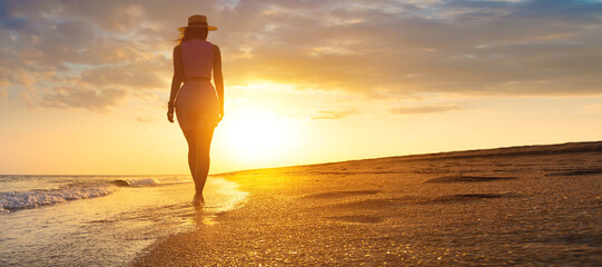 Photo of a young stunning woman walking along the seashore. Fresh sea breeze, a girl watches the sunset and walks towards the sun's rays