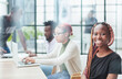 © Katsiaryna - young African American woman in casual clothes posing for the camera while sitting at the table against the background of her colleagues