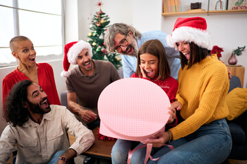 Surprised girl opening Christmas presents at home. Happy and excited family wearing Santa Claus hat gathered together for gift exchange. Positive and joyful domestic relationships in winter vacations.