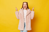 Fototapeta  - Pretty brown haired girl wearing pink fur coat with ponytails isolated over yellow background stands with crossed fingers warm smile emanating strong belief in positive outcomes