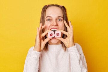 Wall Mural - Funny happy cheerful young girl with ponytails in jumper biting marshmallow posing isolated over yellow background looking at camera eating desserts.