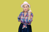 Portrait of mature woman wearing checkered shirt and hat shows middle finger impolite gesture disrespects someone looks rude at camera. Indoor studio shot isolated on yellow background.