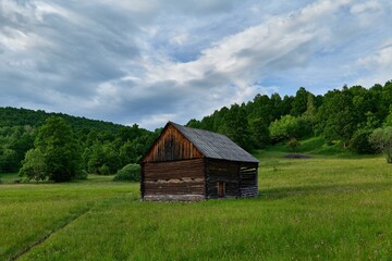 Poster - Aged wooden cabin amongst a picturesque landscape of lush green grass and majestic mountain