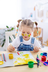 a little child girl paints a drawing in an album and smiles with a dirty face and hands, a happy child draws a picture at home at the table