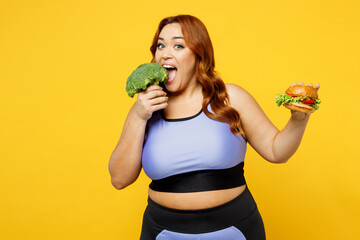 Young happy satisfied chubby overweight plus size big fat fit woman wear blue top warm up train eat bite broccoli hold burger isolated on plain yellow background studio home gym Workout sport concept