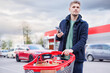 © Girts - A man with a full grocery cart uses his mobile phone in a supermarket, capturing the digital age lifestyle of grocery shopping. The cart holds a variety of items, set against a red and grey backdrop