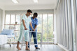 © amorn - Young female nurse help patient man walk with walker careful at hospital. African American female nurse caring patient man walking with walker