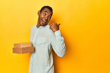 African American man with food boxes, yellow studio, points with thumb finger away, laughing and carefree.