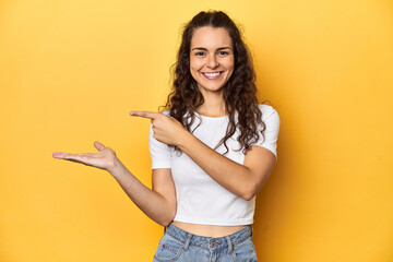Poster - Young Caucasian woman, yellow studio background, excited holding a copy space on palm.