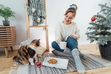 Young Woman In Cosy Sweater With Cat Pet, Drinking Hot Tea, Relaxing On Floor Cushions Near Potted Christmas Tree In Modern Scandi Interior Home. Eco-friendly Cozy Winter Holidays. Selective Focus