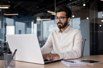 Serious young Indian businessman man working focused on laptop while sitting in office at desk