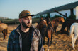 A young farmer near a herd of horses on a farm. Portrait of a bearded young man against the background of horses at sunset.