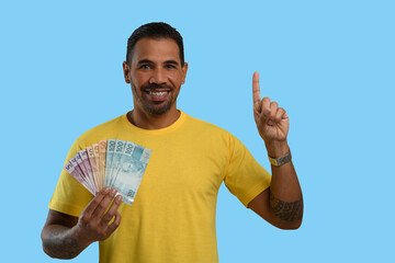 A man with Brazilian money in his hand points upwards with his finger to an empty space on the right-hand side of the picture, yellow clothes, blue background