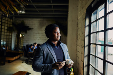 Young African American businessman using his smartphone and enjoying a cup of coffee on a break in a startup company office