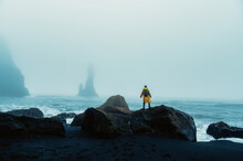 A Man In A Raincoat Is Standing On A Rock And Looking At The Stormy Sky And Waves In Iceland.