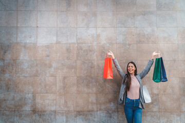 Beautiful smiling young woman standing outdoors in front of monochromatic wall and holding shopping paper bags high in the air. Satisfied young woman celebrating after successful shopping.