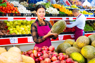 Sticker - Asian woman in uniform standing in greengrocer and holding melon in hands. Her male colleague working in background.