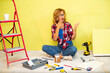 A young beautiful woman in safety glasses, a red checkered shirt, jeans and white sneakers sits on the floor where there are a lot of tools and looks at her stained hands in white paint.