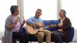 © kaew6566 - Community positive healthy retired elderly playing guitar music song relaxation laughing sitting together on sofa. Elderly man play guitar with friends enjoy leisure entertainment music hobby at home.